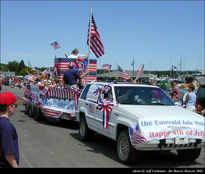A flag-covered SUV and trailer carry people in a parade beside a waterfront lined with boats.