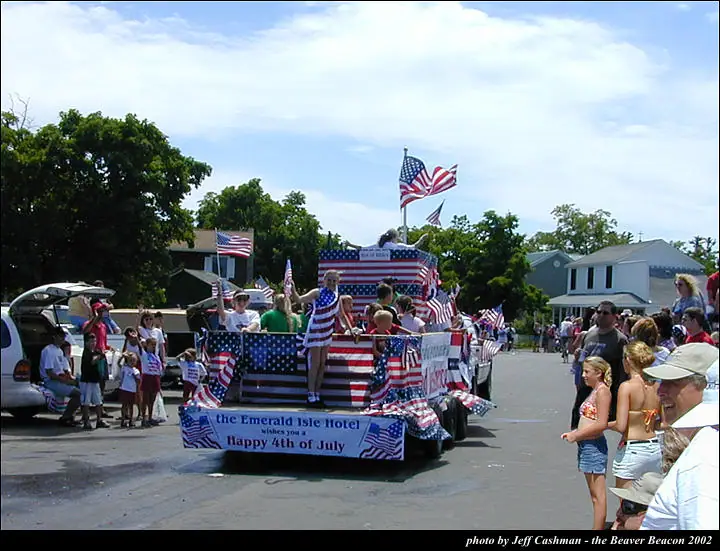 A flag-covered parade float with people aboard moves past spectators on a residential street.