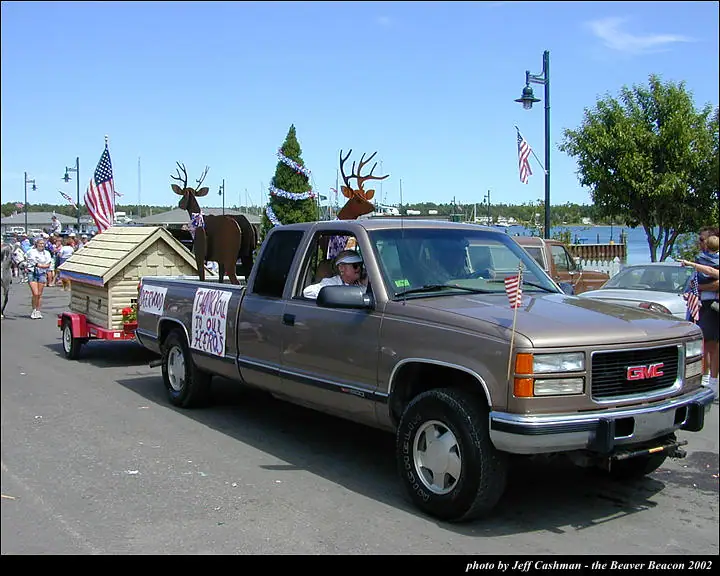 Decorated pickup truck towing a doghouse-like float past spectators and a waterfront during a parade.