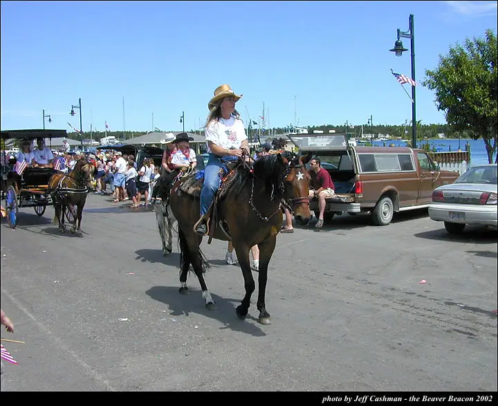 A woman rides a dark horse past a horse-drawn carriage and spectators along a waterfront street.