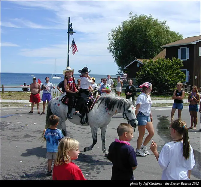 Children ride a decorated gray horse past spectators along a waterfront street during a parade.