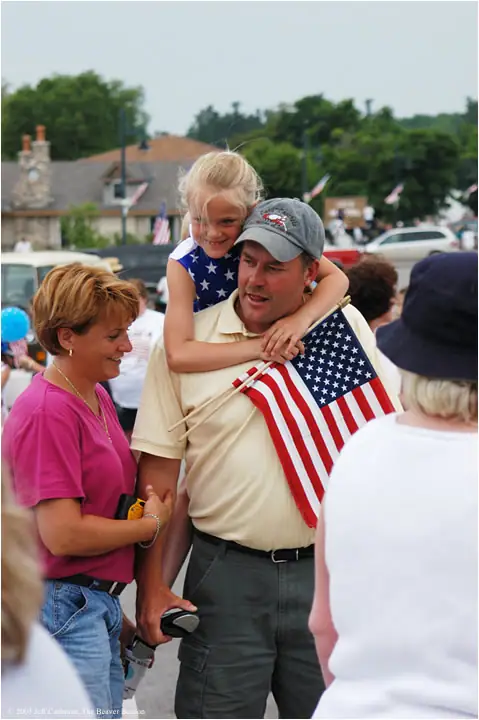 An adult carries a smiling child in a flag-themed shirt through a crowd beside a woman in pink.