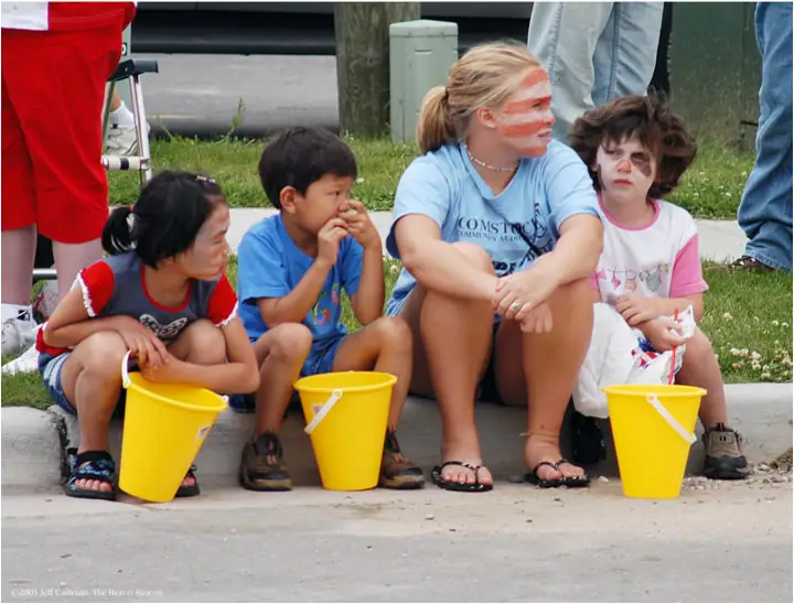 Three children and an older youth sit with yellow buckets while wearing face paint outdoors.