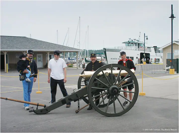 People in historical-style uniforms pose with a field cannon beside a ferry and sailboats.