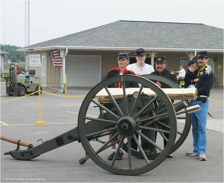 Adults and a child in military-style uniforms pose behind a large wheeled cannon outside a building.