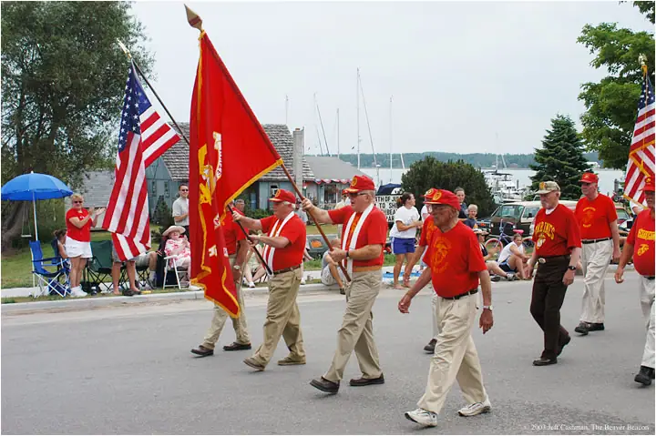 Adults in red shirts march with American and Marine Corps flags along a waterfront road.