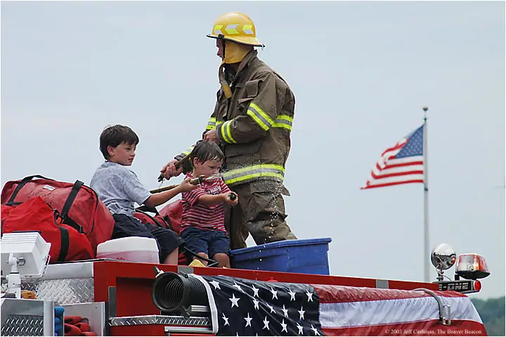 Firefighter and two boys ride on a decorated fire truck beneath an American flag.