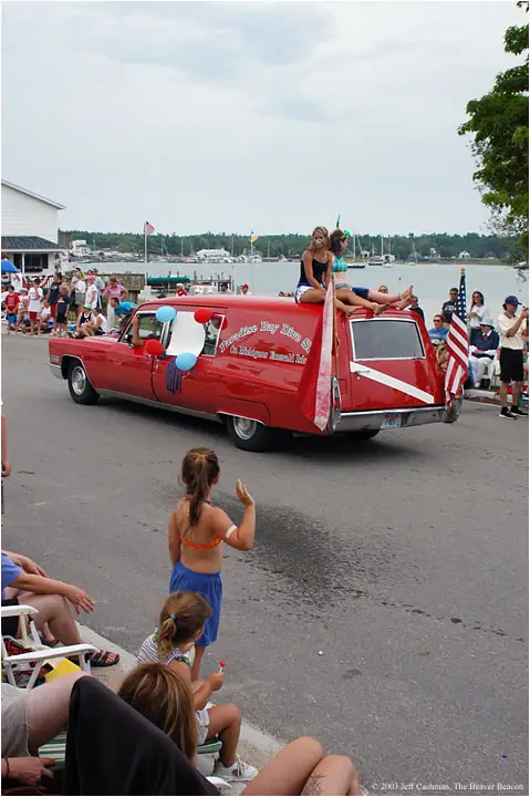 A decorated red vintage vehicle carries two women past spectators along a waterfront street.