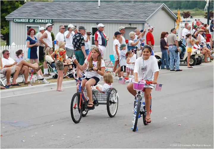Children ride decorated bicycles past spectators along a street during a parade.