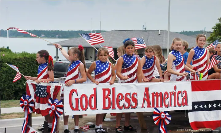 Children in patriotic costumes stand behind a red, white, and blue banner reading “God Bless America.”