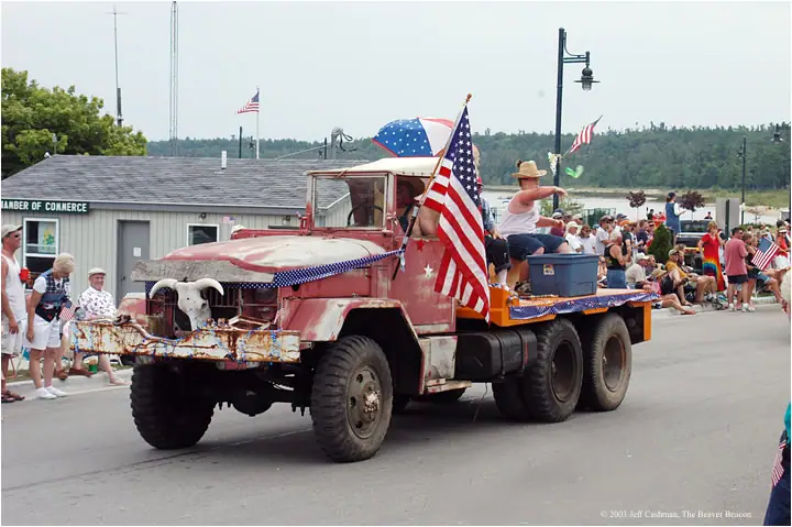 Decorated red truck carrying people passes a roadside crowd during a patriotic parade.