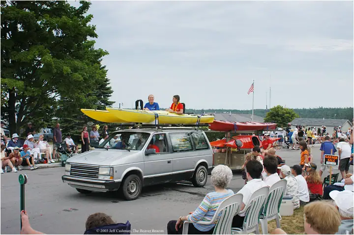 A silver minivan carries kayaks past seated spectators during a parade.