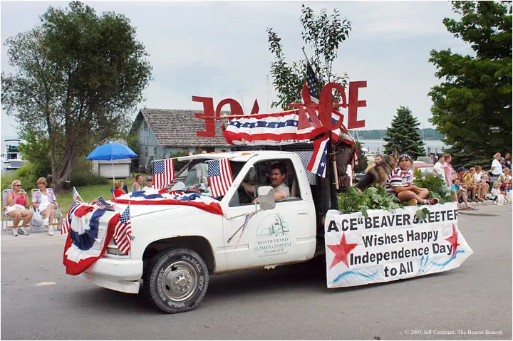 A flag-draped white pickup carrying large red letters and a greeting banner passes spectators in a parade.