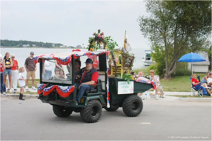 A decorated utility vehicle carries people and flowers along a road beside the water.