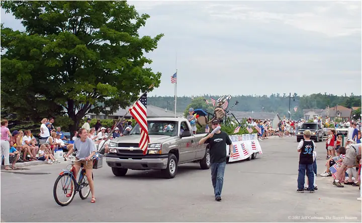 A decorated pickup truck leads a crowded street parade with flags, a bicyclist, and a man using a megaphone.