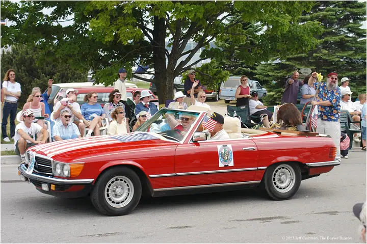 A red convertible decorated with American flags passes a crowd of spectators during a parade.
