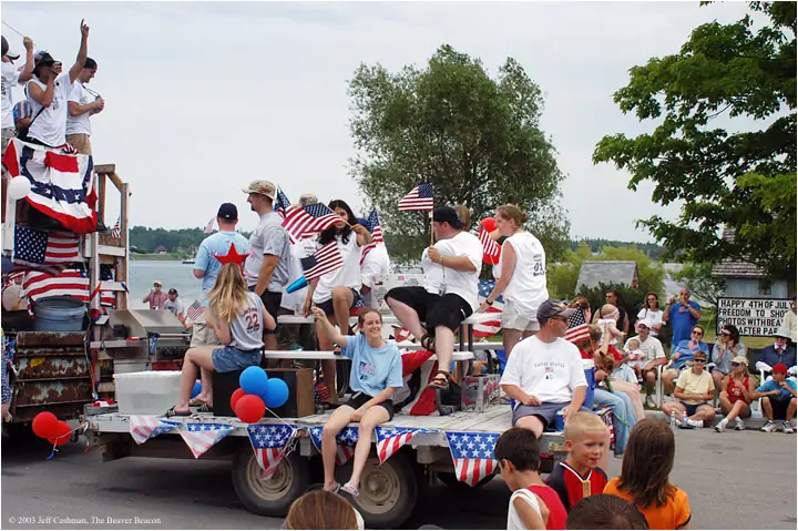People ride a flag-decorated parade trailer while spectators gather along the waterfront.