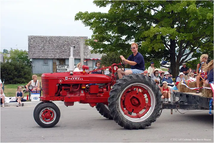 A red Farmall tractor pulls a hay-filled trailer past seated spectators during a parade.