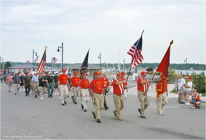 Marchers carrying American and ceremonial flags pass a marina while spectators watch.