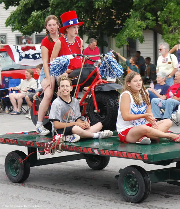 Young people ride a decorated parade float with a red all-terrain vehicle and patriotic decorations.