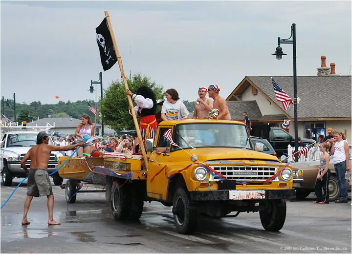 A decorated yellow truck and trailer carry costumed participants while a person sprays water beside the street.