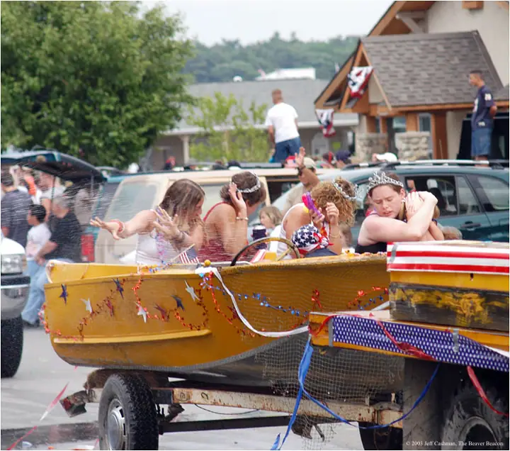 Women in tiaras ride a flag-decorated boat trailer during a parade.