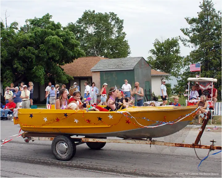 A decorated yellow boat carrying people rolls on a trailer past a crowd during a parade.