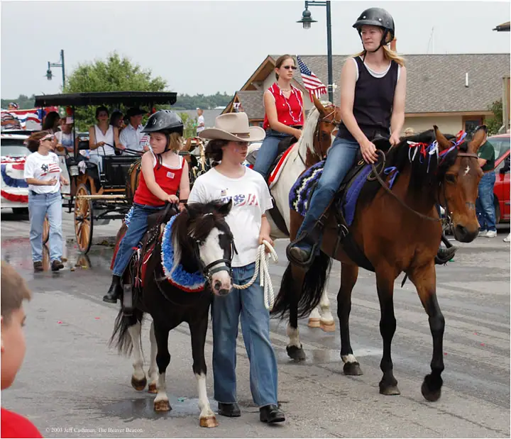 Horse riders and a horse-drawn wagon proceed along a decorated street in a parade.