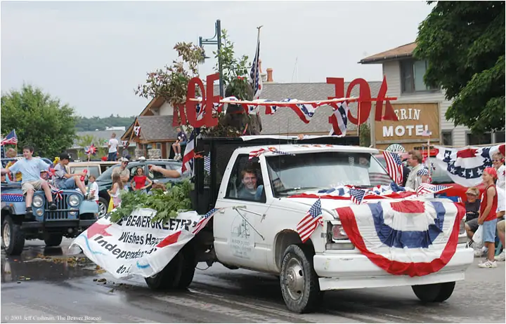 A flag-draped white pickup truck leads a decorated vehicle parade along a street.