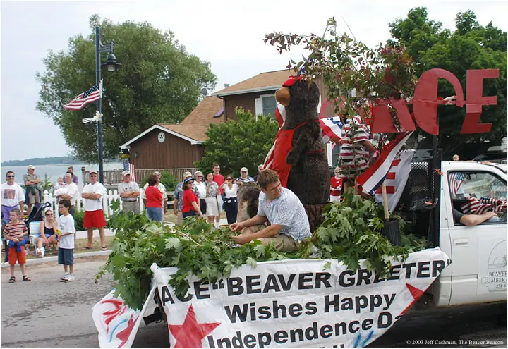 A parade float with a beaver mascot, patriotic decorations, and a banner passes a crowd.