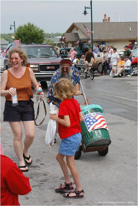 People gather on a street as a bearded man pushes a decorated green barrel cart beside a child.