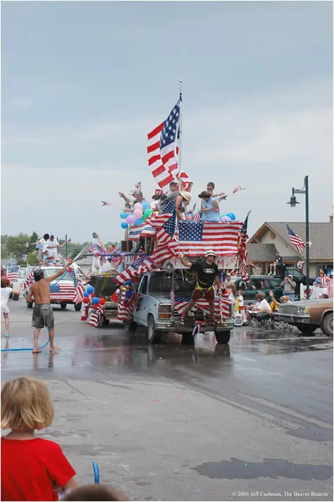A flag-covered parade truck carries people and balloons along a wet street as water sprays across the pavement.
