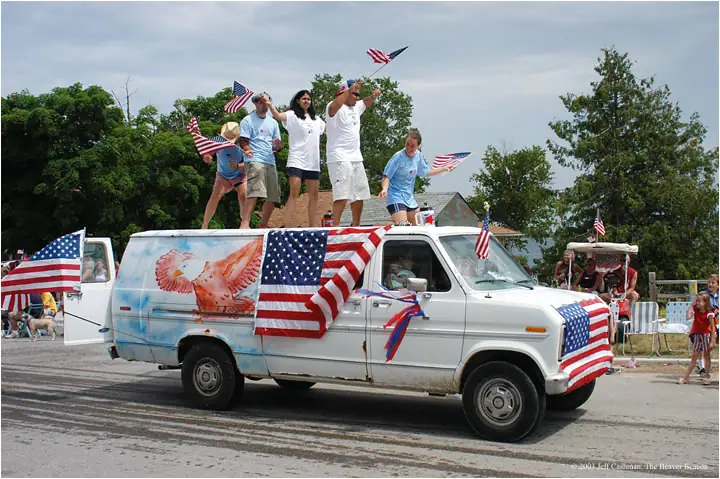 A flag-decorated van carries people waving flags during a parade.