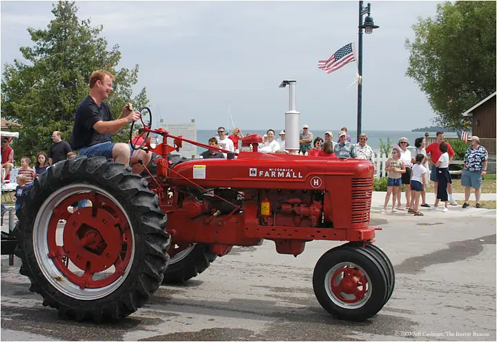 A man drives a red Farmall tractor past spectators gathered along a waterfront road.