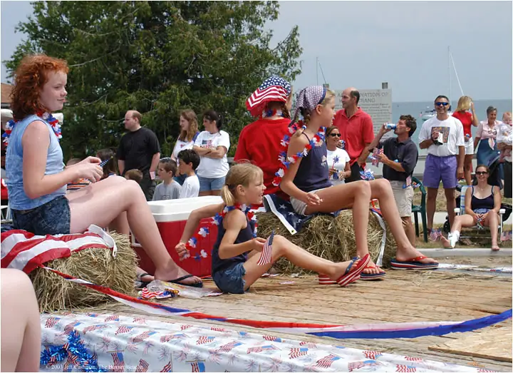 Children and adults in patriotic clothing gather on a decorated wooden platform beside the water.