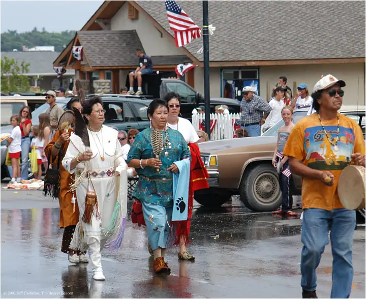 A parade group in fringed regalia walks along a wet street while a drummer leads them.