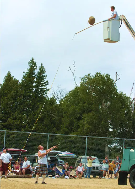 A kilted athlete and spectators watch a suspended object being guided from an elevated work platform.
