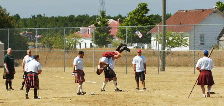 People in kilts take part in an athletic event on a dry field, with houses and a chain-link fence behind them.