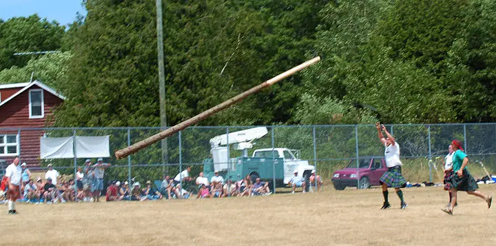 A kilted athlete throws a long wooden pole across a field as spectators watch.