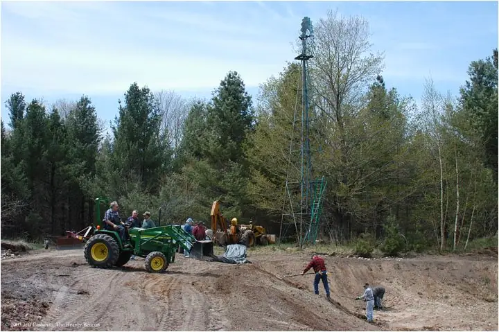 People and heavy equipment work on a cleared dirt site beside a forest, with a tall metal tower in the background.