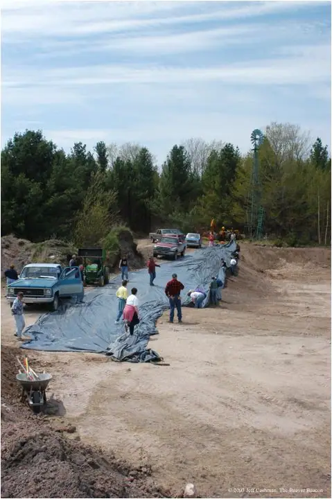 People spread a large plastic liner across an excavated dirt area beside several vehicles and a wooded area.