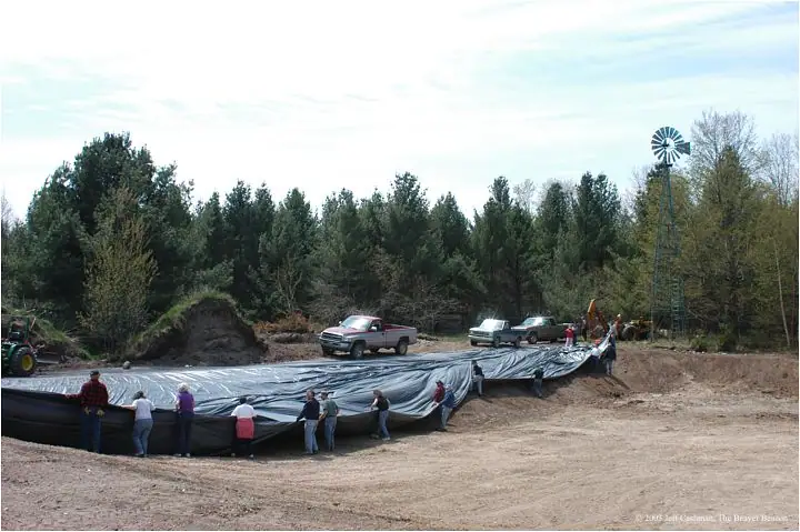 People spread a large black liner across an excavated basin beside trucks, heavy equipment, trees, and a windmill.