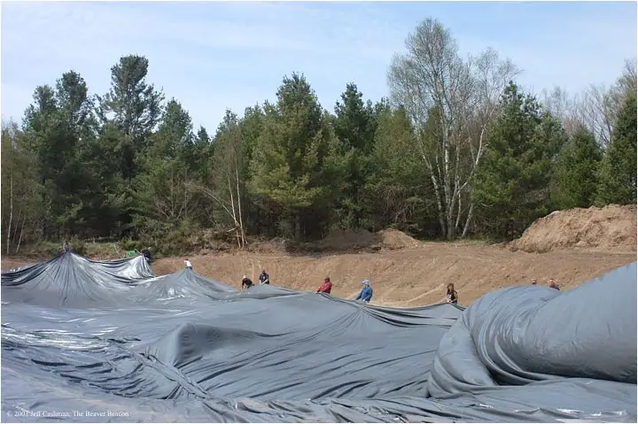 People spread a large gray liner across an excavated area beside a forest.
