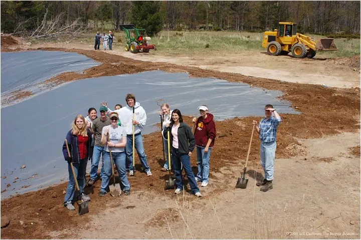 People pose with shovels beside a plastic-lined pond under construction, with a loader and tractor behind them.