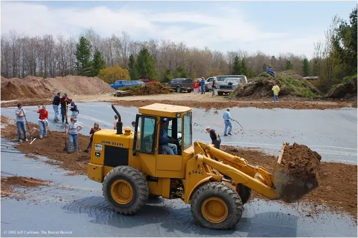 A yellow John Deere loader moves soil across a lined excavation while people work nearby.