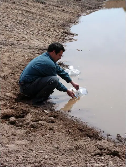 A man crouches beside a muddy pond, handling plastic bags at the water’s edge.