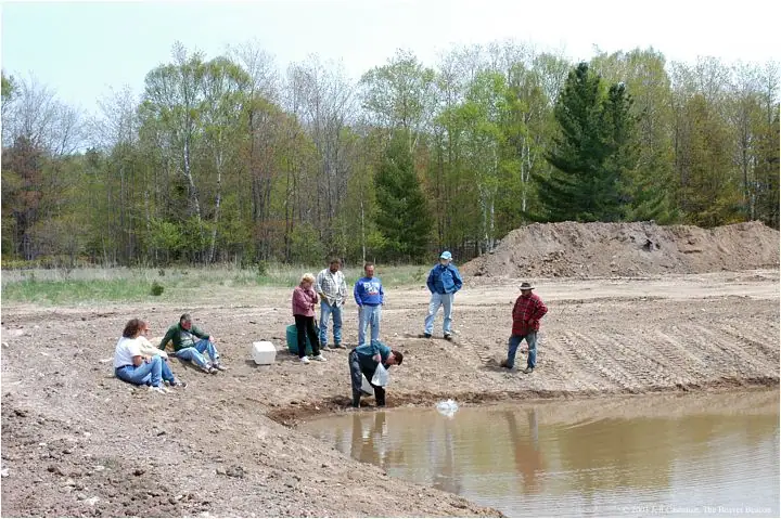 People gather beside a muddy pond under construction, with a forest and large soil pile behind them.