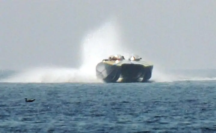A high-speed motorboat with two occupants throws up a broad spray as it moves across the water.