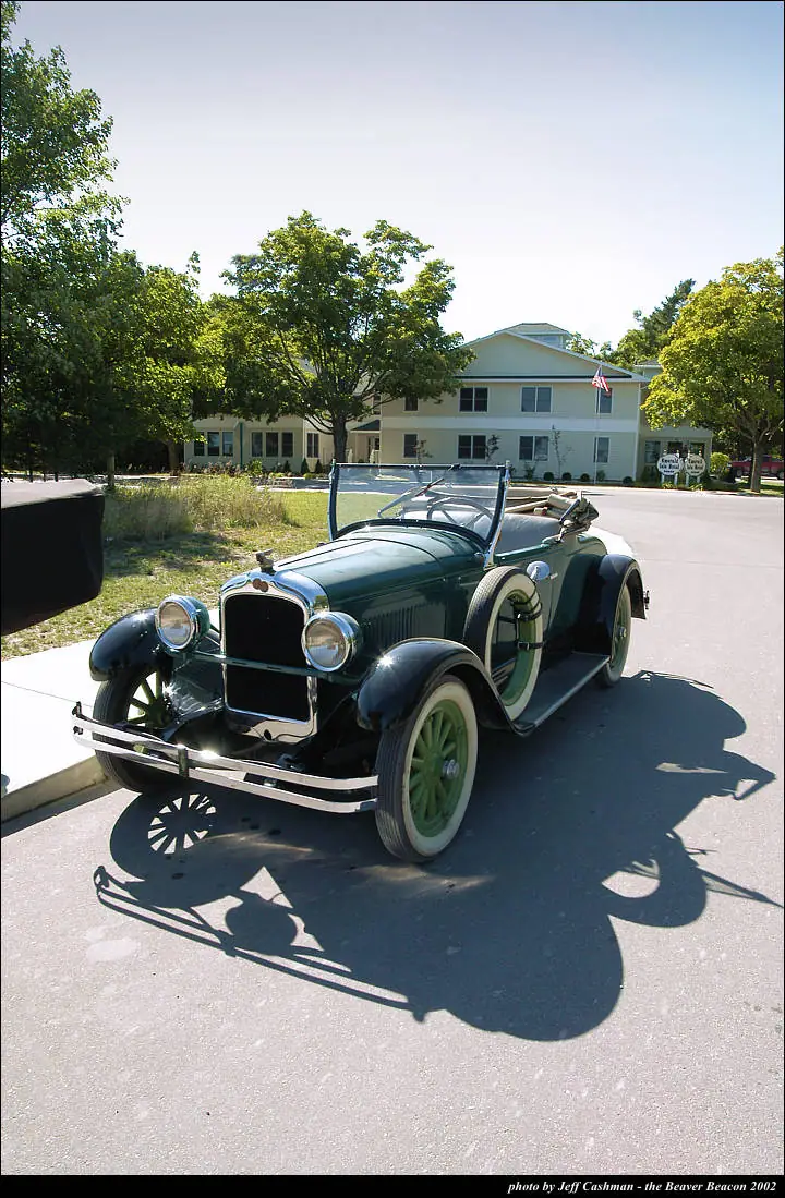 A dark green vintage convertible is parked outside a light-colored building beneath leafy trees.