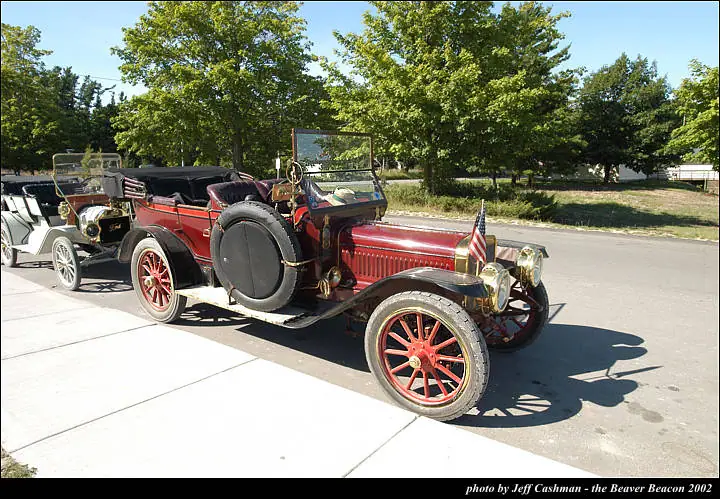 A red antique automobile with a side-mounted spare tire is parked beside other vintage cars on a sunny street.